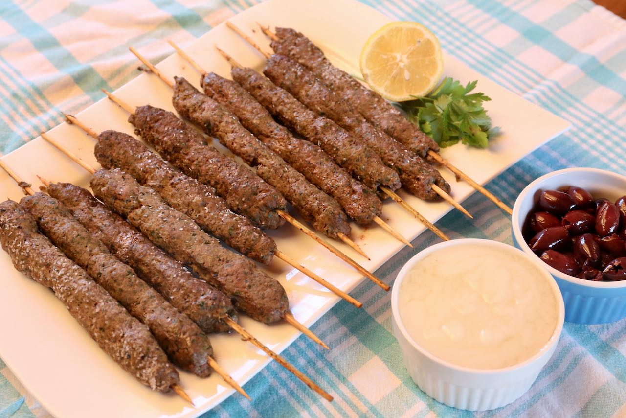 Serve barbecued Afghan Kebab Beef Kafta skewers on a platter with lemon, parsley, toum and olives.